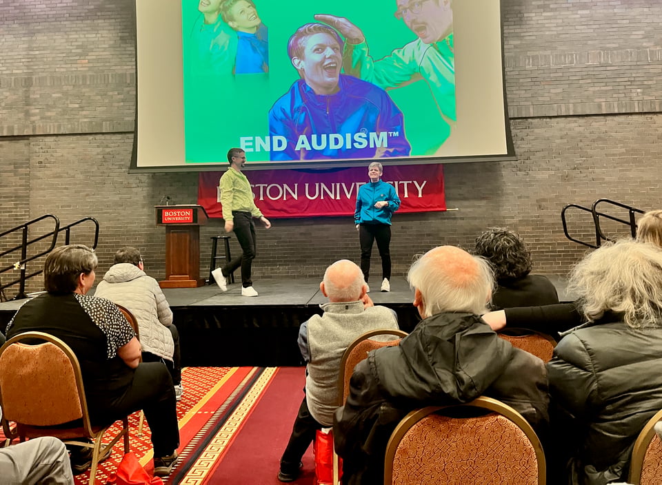 Andrew Bottoms and Megan Malzkun, two white college professors, stand on a stage in front of a large audience wearing bright green and blue windbreakers, respectively. Behind them is a projection showing their faces in various comic poses, with the words END AUDISM™ superimposed. Below the screen is a red banner with BOSTON UNIVERSITY written on it.