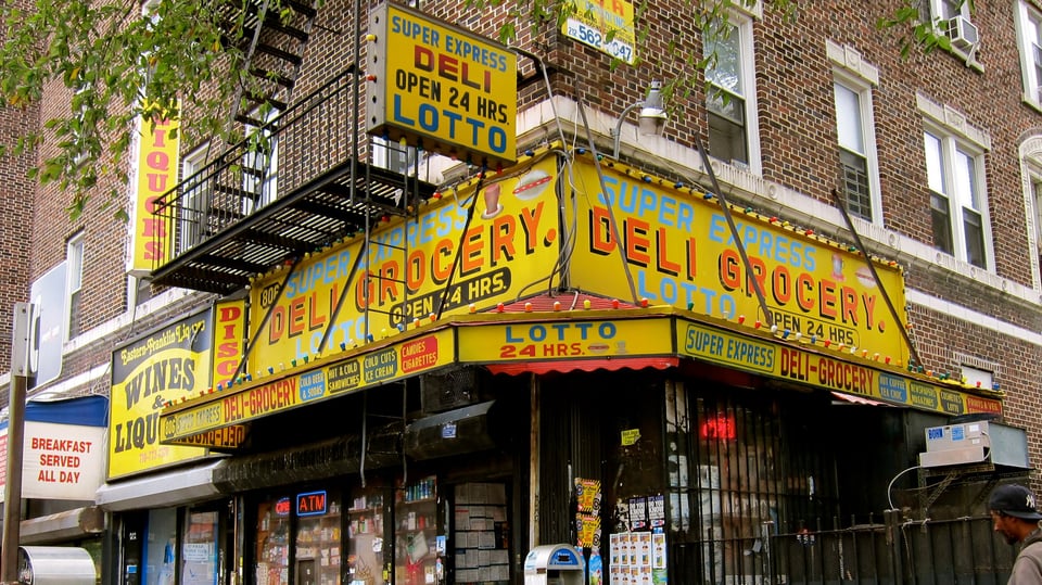 A photo of a deli in New York City, focusing on the sign: it is bright yellow, with bright, red lettering that says "deli grocery" on it. Jutting out from the corner of the store is a hanging sign, ringed by a few lightbulbs (the others are apparently broken and no longer work); the sign is yellow and has text on it saying "super express deli 24 hrs, lotto"