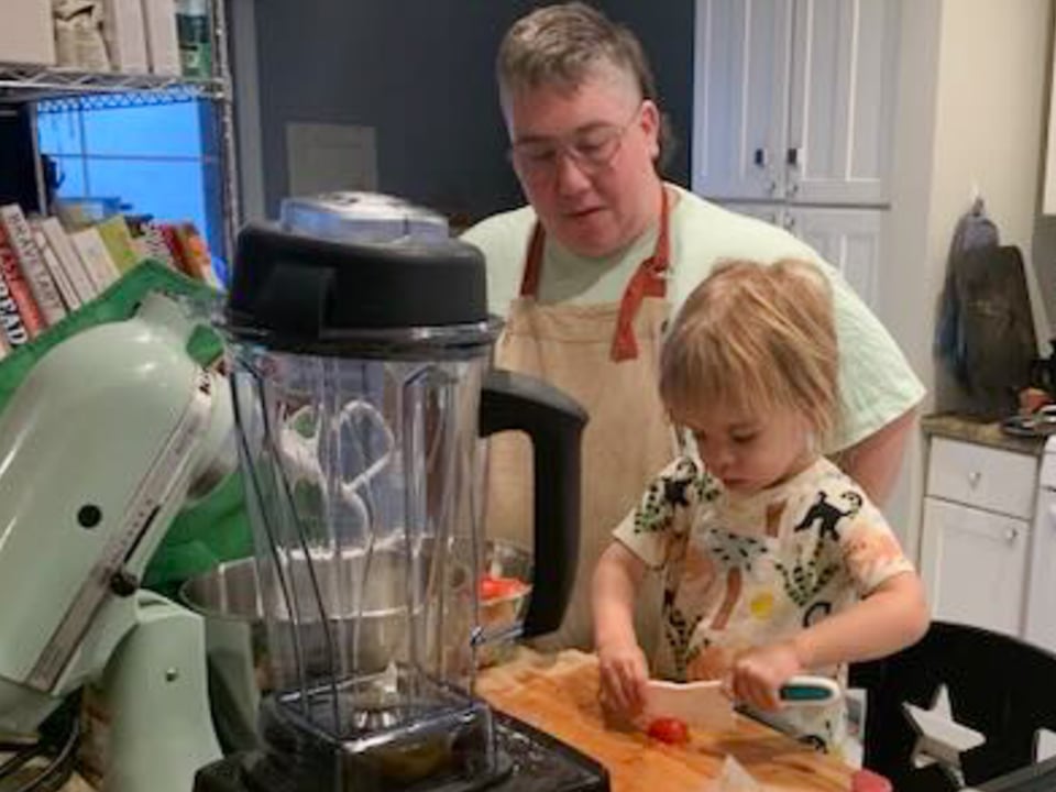 A toddler cuts a red tomato in half on a wooden cutting board with a kids' knife while his parent watches, wearing a beige apron and a green shirt. In the foreground is a mess of kitchen equipment, a blender and a stand mixer