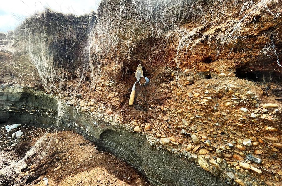 A trowel and a metal cylinder stuck into a wall of rocks and sediment along the bank of a stream