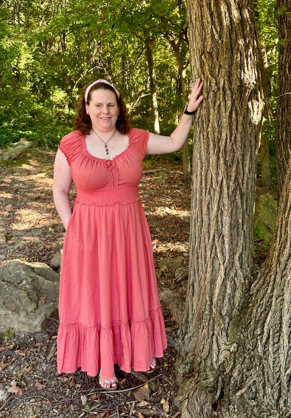 Daisy, a fair-skinned woman with freckles, standing in a wooded area with her left hand resting on a tree and her right hand in her pocket, wearing an ankle-length coral-colored dress with cap sleeves, a scoop neckline with a tie, a cinched waist, a flowing skirt with a ruffled hem, and little textured dots all over, a shiny pink fabric headband holding back her auburn curls, a necklace with a vial of flower petals and a dangling flower charm, pink flower earrings with stones in the center, and tan strappy sandals with 3D flowers on the top that may or may not be visible