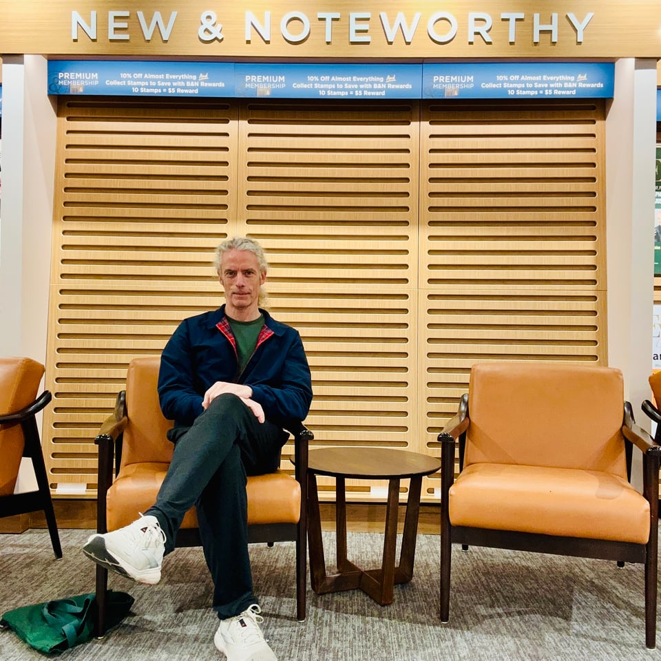 Photo of white man in navy Harrington G9 jacket, green pullover, black sweatpants & white sneakers. He is seated in front of an empty bookstore wall with the words NEW & NOTEWORTHY above him