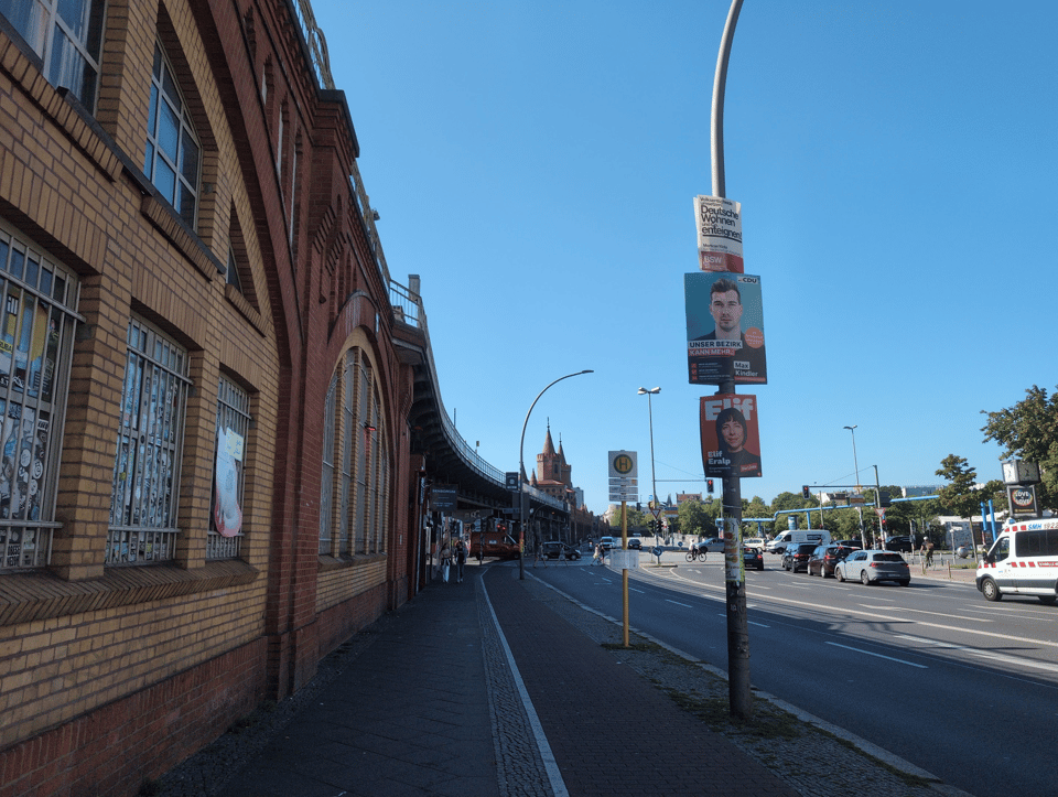 A view from near the bottom of the Warschauer Str. hill toward the Oberbaumbrücke, seen as two pointy towers in the background. In the foreground (left) is the brick building that is also the train bridge, which curves slightly to the right as it approaches the river/background. The angle of the photo combined with the curve makes the perspective slightly fish-eye. In the mid-foreground (right) are three election posters on a light pole. They are for the BSW, CDU, and Linke.
