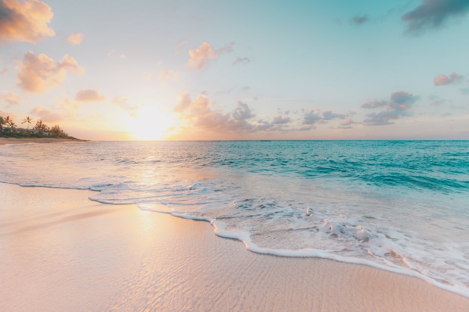 A photo of a sunset on the beach in Oahu, Hawaii. There is pale, clean sand in the foreground and blue water in the background.
