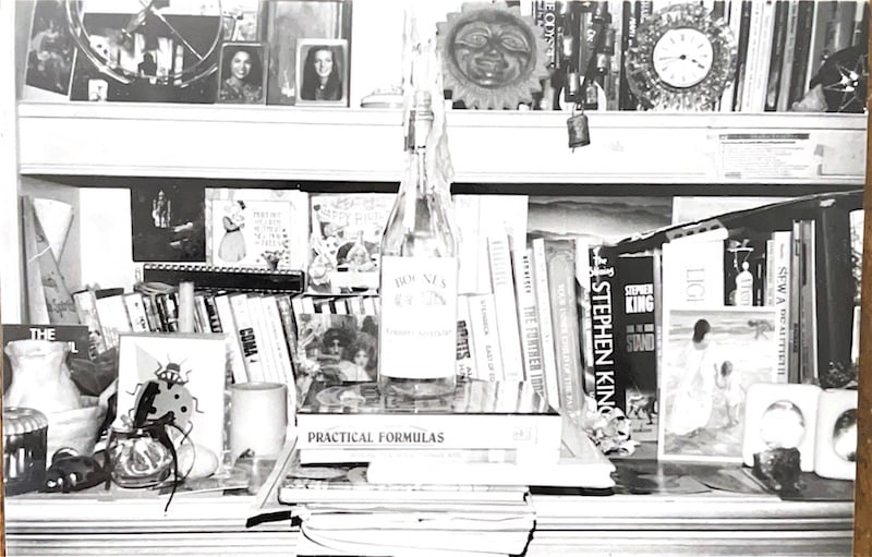 a black and white photo of a chaotic desk with bookshelves. Numerous books by Stephen King, an empty Boone's bottle atop a stack of books with old wax dripped over its sides. Numerous tchotchkes and postcards. 