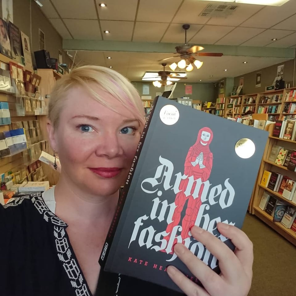 Kate has floppy assymmetrical hair and is holding a copy of Armed in Her Fashion.