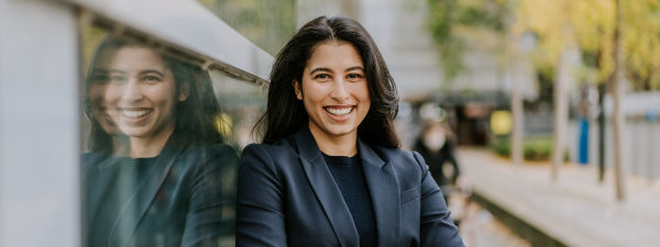 A picture of Seham Areff. She is at the centre of the image, wearing a grey blazer and black t shirt. She has dark hair and is smiling with her teeth.