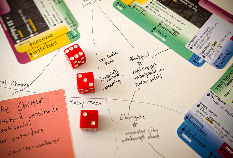 Close-up of a game of Downcrawl in progress, showing 6-sided dice, a map with evocative locations like "Bleakport" and "Ebongate" written on it, and a selection of Downcrawl Deck cards with prompts like "serene witches" and "heartless diplomat."