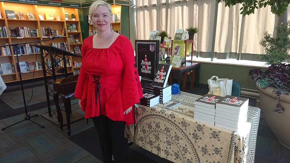 Kate in her red medieval tunic by the book table. The table has copies of Armed in Her Fashion.