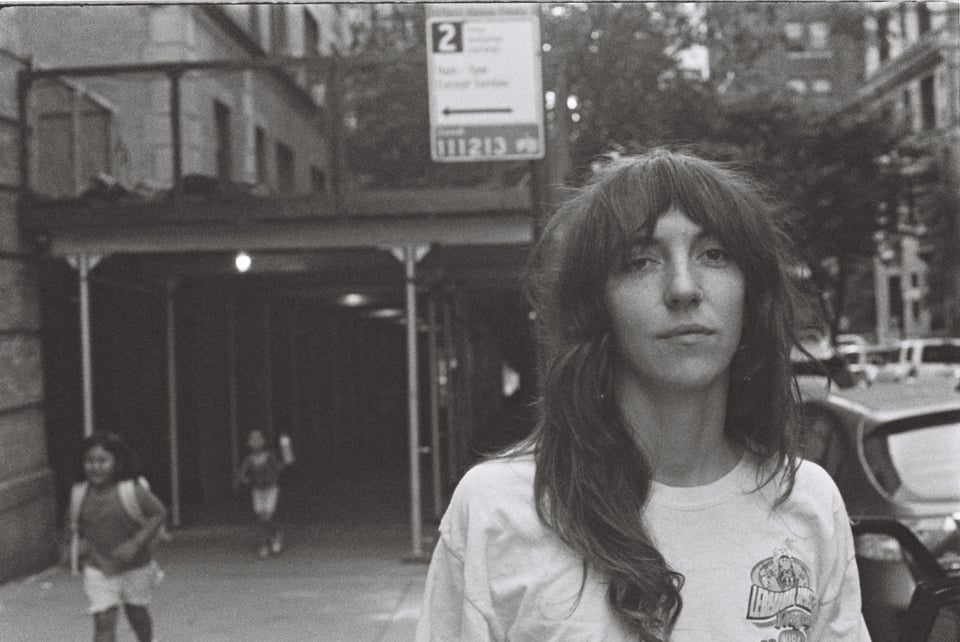A black and white image of a woman with hair parted in the middle and falling past her shoulders wearing a white t-shirt on a sidewalk in what looks like New York..