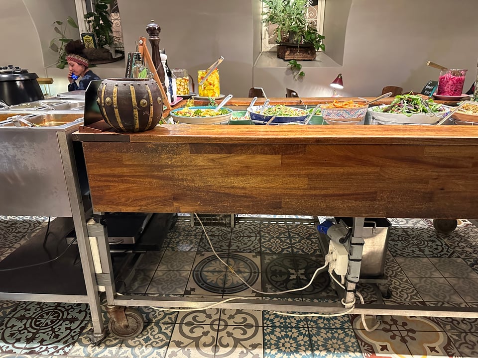 A section of a buffet. Wooden table, with metal cabinet to left. Multiple bowls containing food, plus various jars.