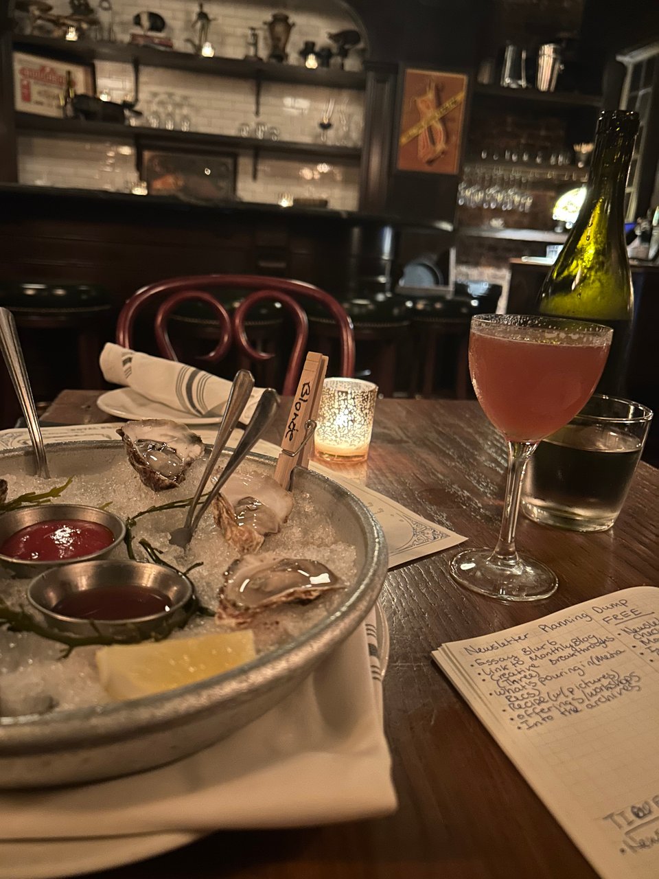 a photograph of a restaurant spread at a table including raw oysters with lemon, a small candle, a pink cocktail, and an open notebook.