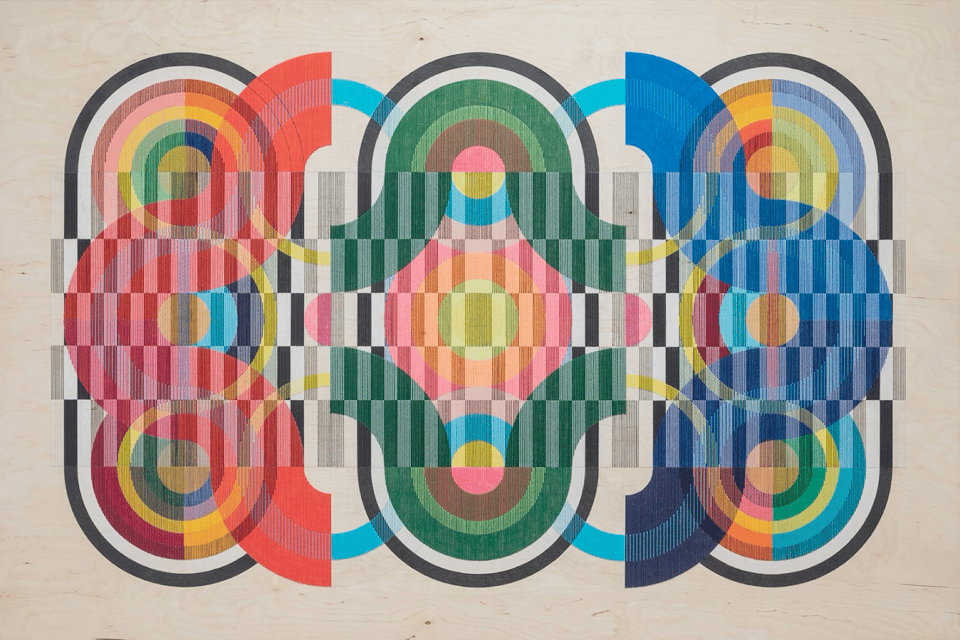 Abstract geometric artwork featuring symmetrical patterns with vibrant overlapping circles and semicircles in various colors, including red, blue, green, yellow, orange, and pink. The composition includes precise vertical stripes and grids intersecting with circular forms, creating an intricate visual effect on a light wooden background