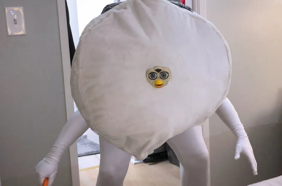 A human in a body suit wearing a loaf-like sphere with a Furby face