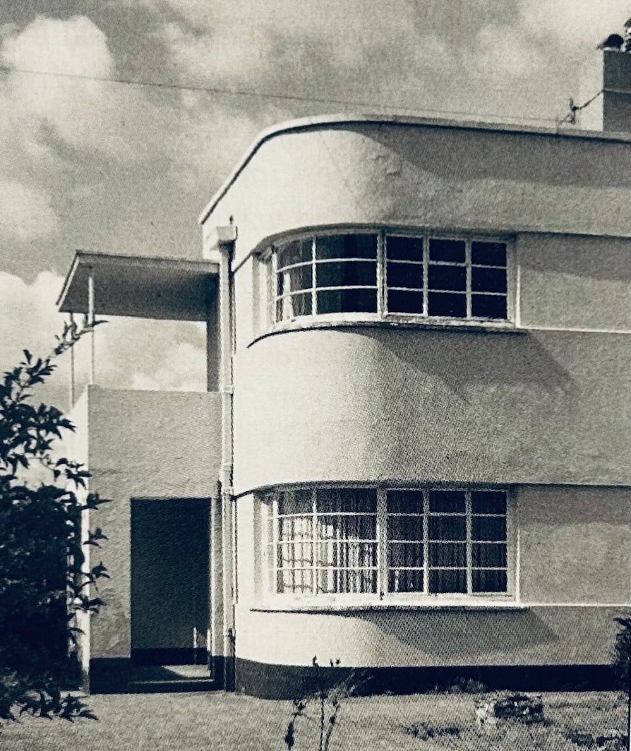 A two storey 1930s house. A curving wall has large curving windows set in it, with metal frames. To one side, a squared off porch is topped by a covered but open balcony