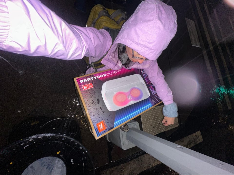 A selfie of a soaked, raincoat-clad Maya holding a packaged speaker.