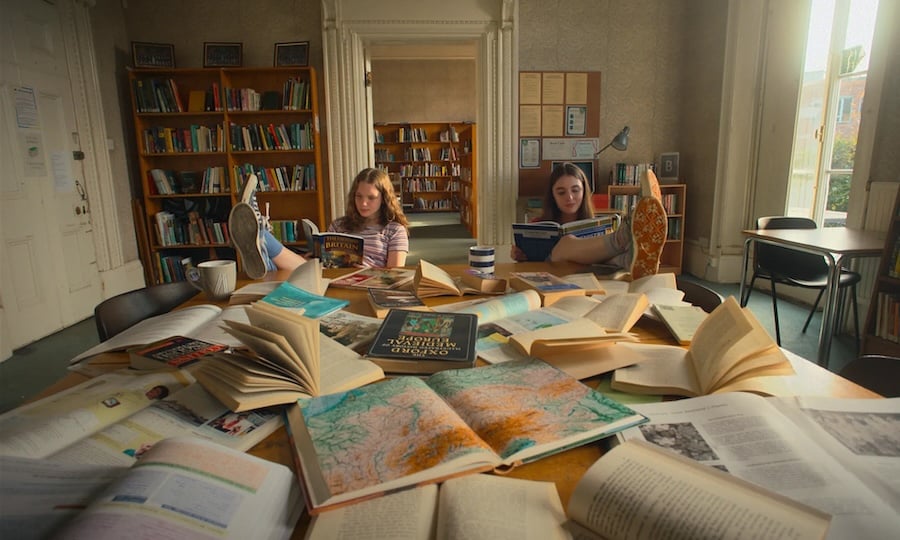 A still from a film: a library with two teenage girls sitting at a big pile of books, each reading a book and with her feet up on the desk