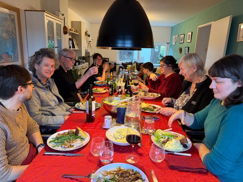 A large dining room table is surrounded by people talking and eating Thanksgiving meal.