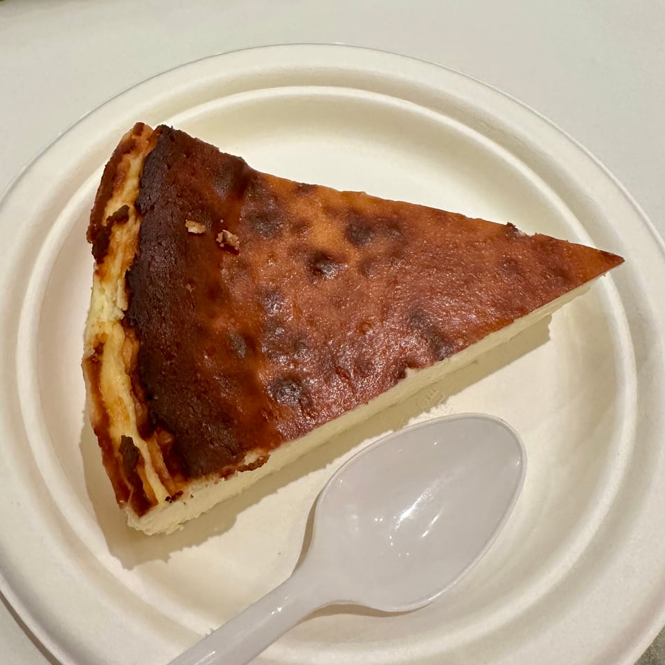 Photo of a slice of Basque Cheesecake, on a paper plate, beside a plastic spoon