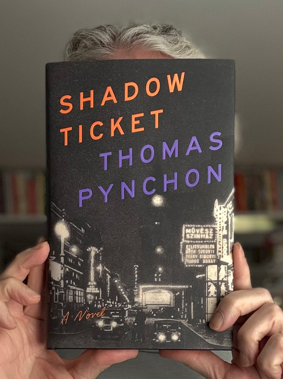 photo of white man holding a copy of Thomas Pynchon's 2025 novel, Shadow Ticket, in front of his face