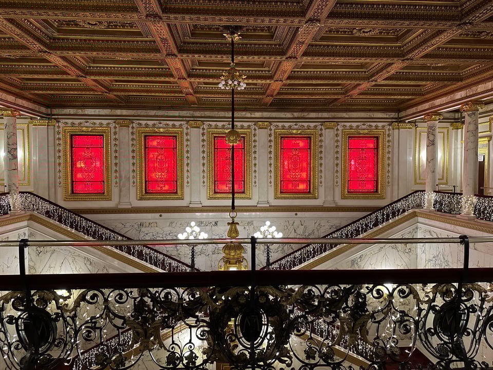 photo above lobby of the Metropolitan Club in NYC, with 5 red-lit windows in the background, stamped metal ceiling, and ornate railing; two sets of stairs along the back wall form an X