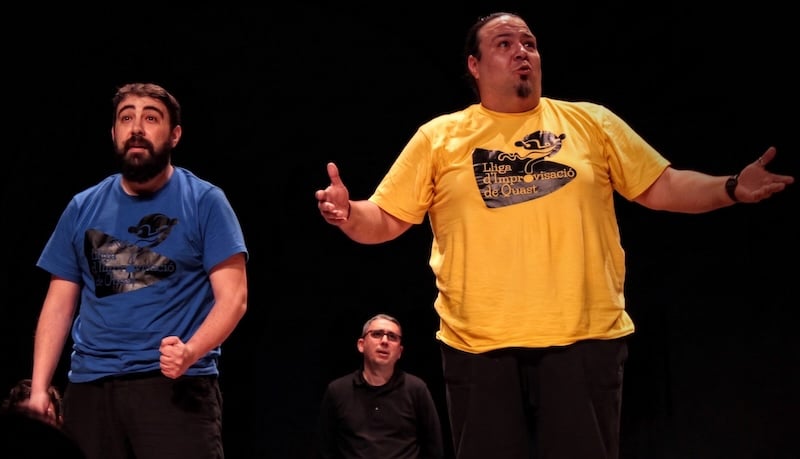 Three performers on stage, with two in colorful shirts expressing emotions through gestures while a third stands behind them.