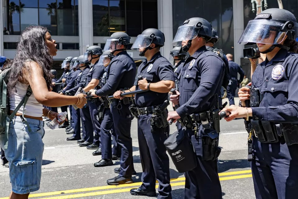 A May Day participant to the left of the frame, wearing a t-shirt, jean shorts, and a backpack, faces a line of 10 plus LAPD officers, wearing riot helmets with shields down, and holding their batons in front of them, ready to strike.