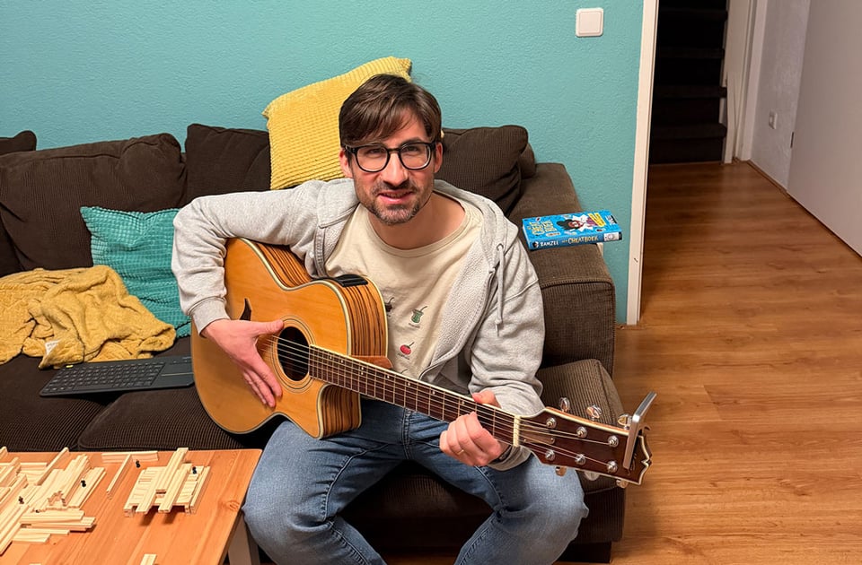 A man smiles at the camera as he sits on a couch and holds a guitar.