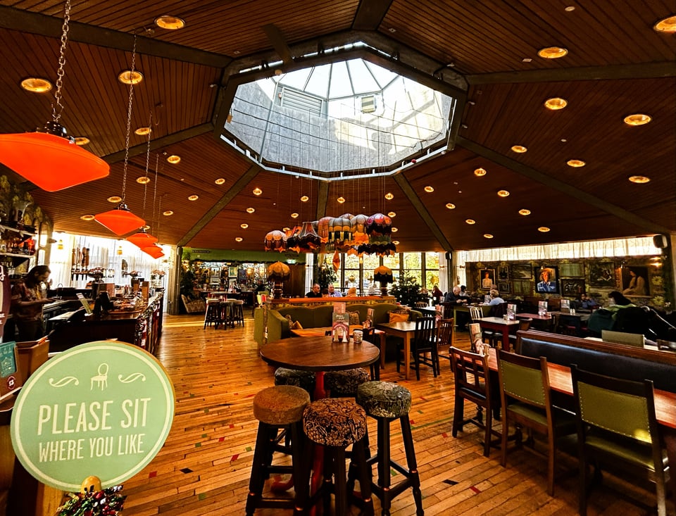 Colour photo of an octagonal interior space. The open plan space has been filled with mismatched vintage furniture. But the real focus is of the wood-lined ceiling and the octagonal concrete ring that holds it in place. Above the ring is an equally octagonal lantern that is letting in light. Suspended below the lantern are scores of orange lampshades with fringing.