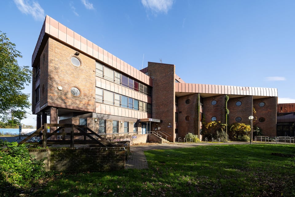 A brick clad office building. It appears to be built in a Y shape. One wing has flat walls with both ribbon windows and portholes. The other wing is made up of squares that have been offset to create a zigzag facade with square windows in it. Both wings have a deep copper clad roof.