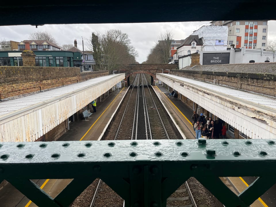 Looking over the edge of a metal bridge, down on two train tracks going through a built up area. Two platforms are on either side of the trains, with a white wooden overhang, and a road bridge further back in the photo.