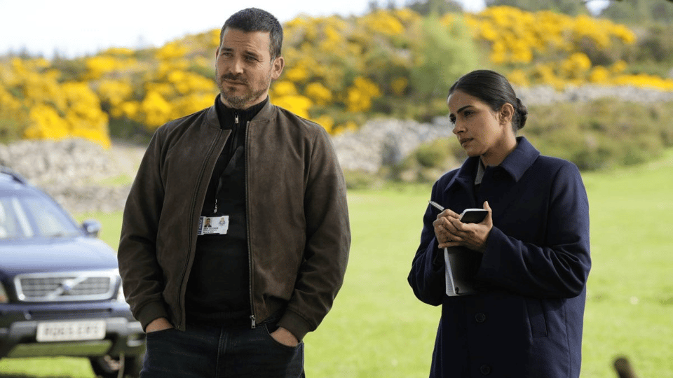 A man and a woman stand in a field, the man wearing a police lanyard and the woman taking notes in a notebook.