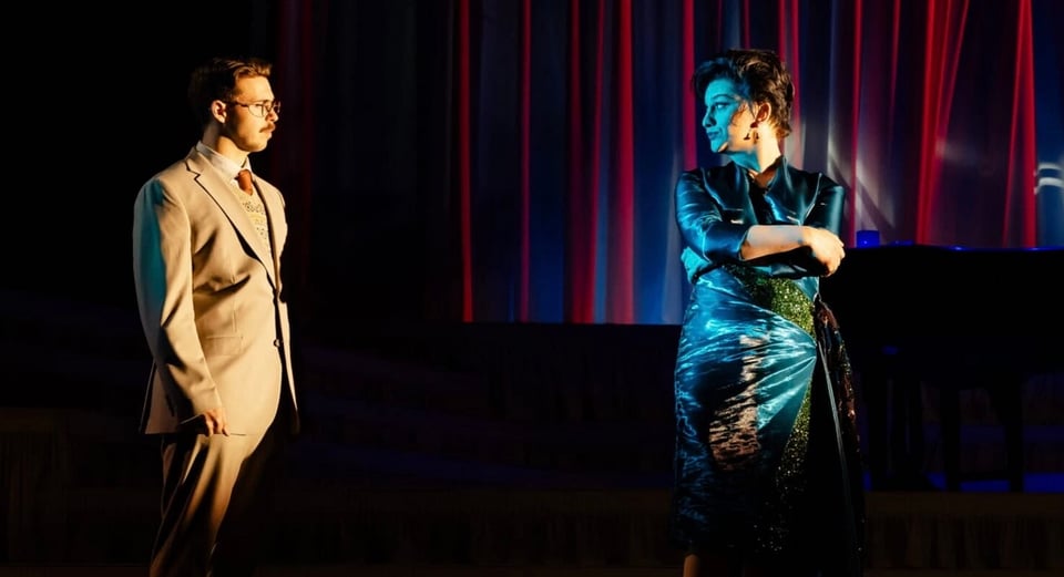 A man in a light-colored suit stands on stage while a woman in an elegant dress poses dramatically, illuminated by colorful stage lights.
