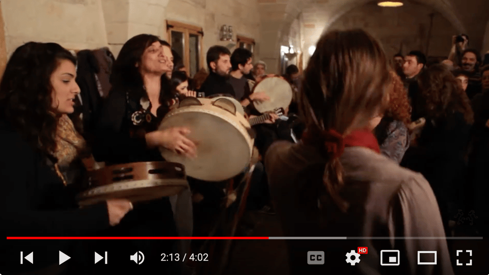 A still from a video of many people playing tamburelli and dancing in a small room