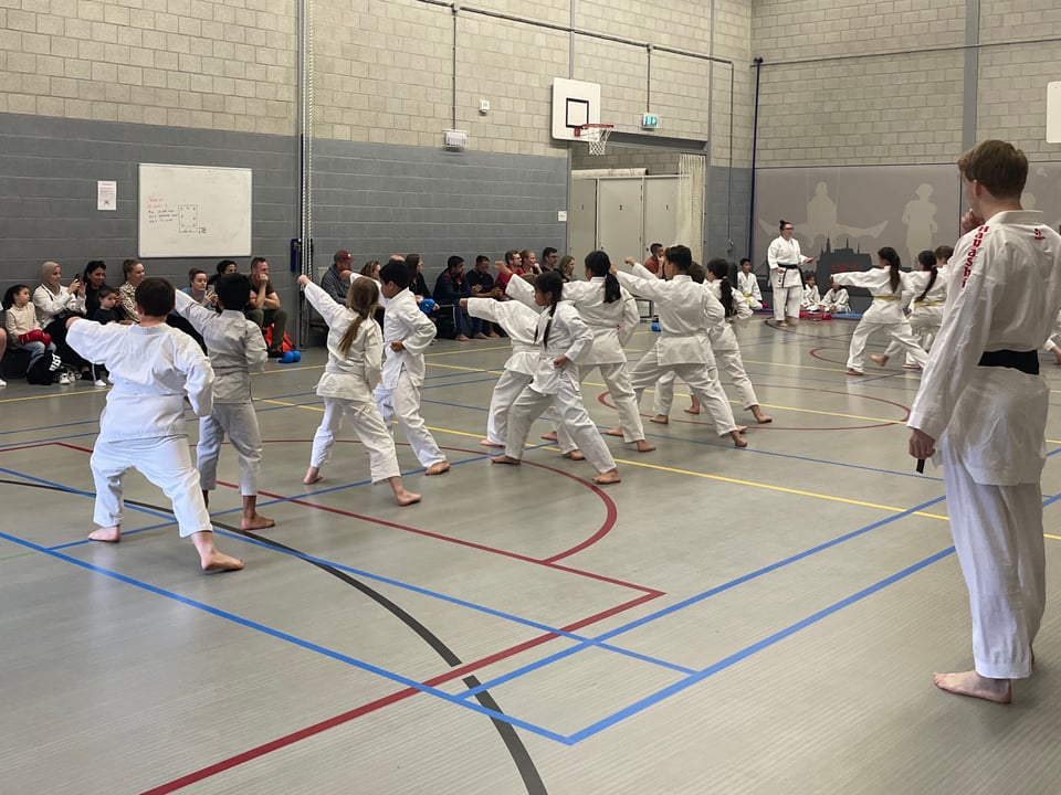 In a grey gym, children in white karate suits all face away from the camera with their left arms outstretched and their right legs behind them. Parents are sitting on benches against the wall watching their children take their exams.