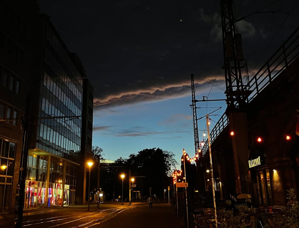 A view of the street just after sunset opening to a sky filled with a rolling wave of grey clouds. (It really looks like a wave). There are yellow street lights, with tram lines on the street, a glass building on the left and an elevated train line structure on the right