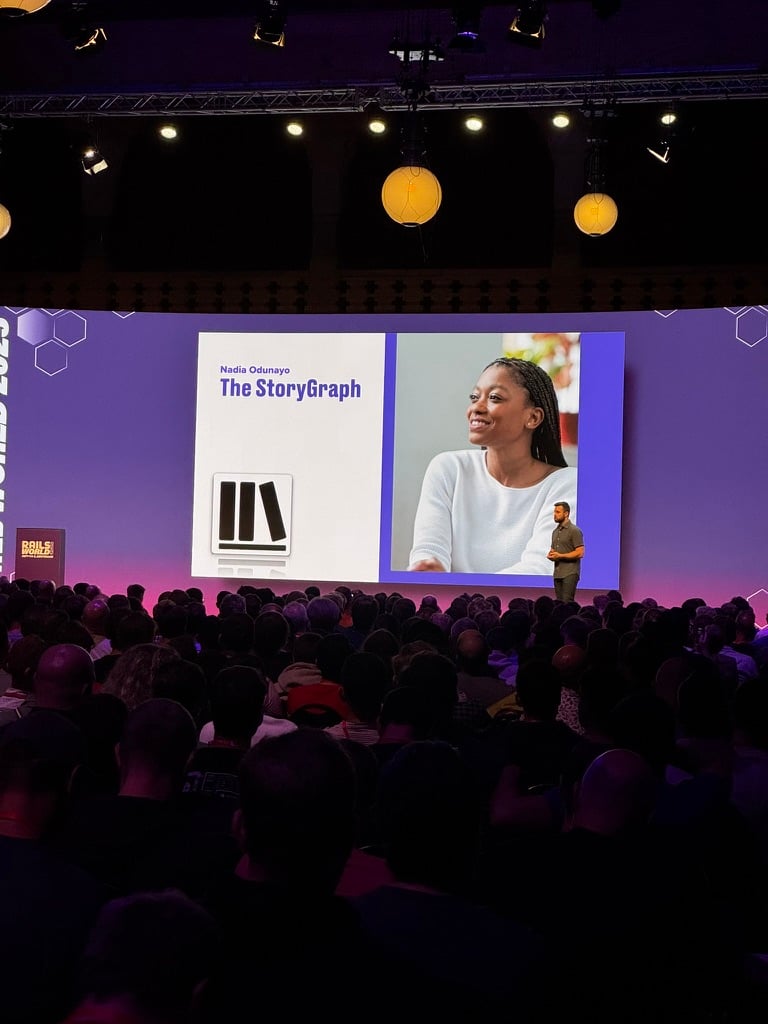 A crowded conference hall with a purple-lit stage. On the large screen, there’s a slide showing a photo of me smiling in a white top, alongside the text “Nadia Odunayo, The StoryGraph” and the StoryGraph logo. Joe is on stage presenting, standing to the right of the screen, while the audience watches attentively.