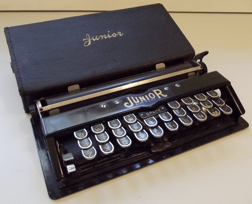 A photograph of a small, mechanical device that is, at a glance, a miniature typewriter. The brand name is "Junior", and it is entirely black. It is perhaps 8 inches wide, 4 inches deep, and an inch tall. The keyboard is very cramped - notably, the space bar is exceptionally small and located at the top center of the keyboard, instead of running along the bottom as with a regular typewriter.