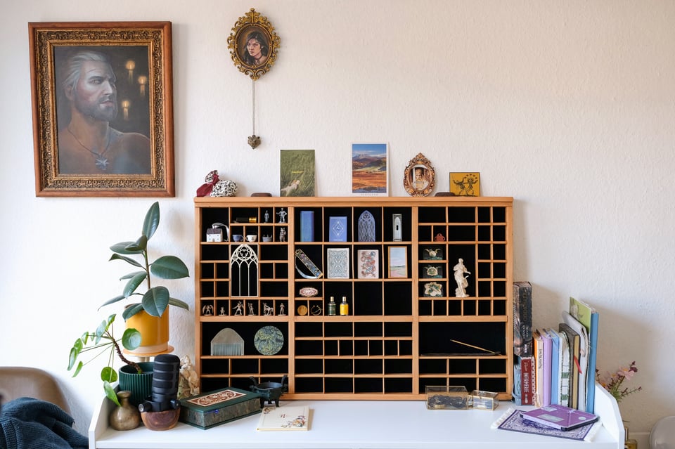Photo of the trinket shelf sitting on a white dresser. On its left are potted plants and on its left are books. There is a large portrait hanging on the wall to the side, and a smaller round portrait above the shelf. The shelf is about half-filled with various colourful small objects.