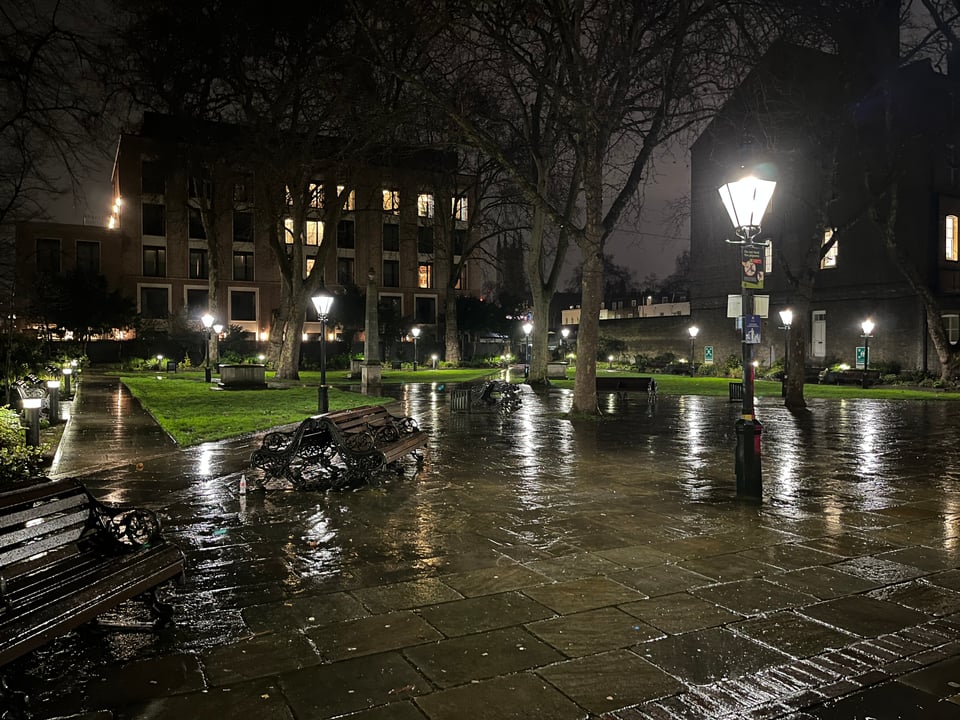 A London park at night, lit up by old-fashioned looking streetlights. It’s rained recently and the pavement is reflecting the water and the light.