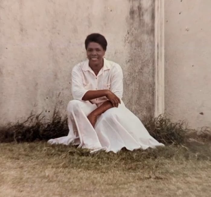 my grandma kneels in front of a wall wearing a white, collared, full-length dress. her elbow is propped on one of her knees so that her arm crosses her body. she smiles.