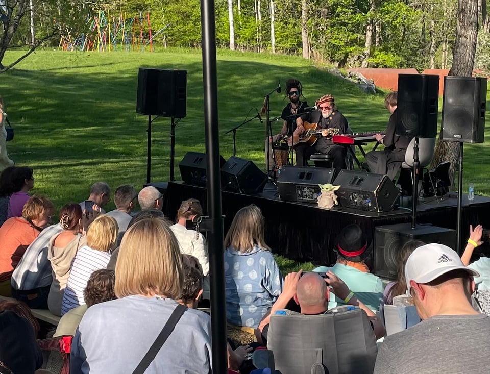 Photo shows Russian rock legend Boris Grebenshchikov on a small stage in a green park, flanked by percussionist Gleb Grebenshchikov and keyboard player Konstantin Tumanov (who is parially obscured). There are the backs of many fans in front, and a weird pole crossing vertically through the center of the image (sorry!). A Baby Yoda doll sits on the edge of the stage.