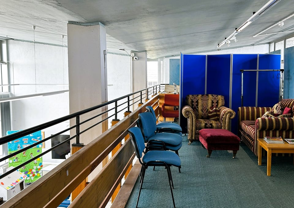 Looking down a gallery, with the drop to the main library space to the left. A mixture of stacking chairs and an old three piece sofa are arranged in front of some old display boards. The internal V created by the butterfly roof is clearly visible.