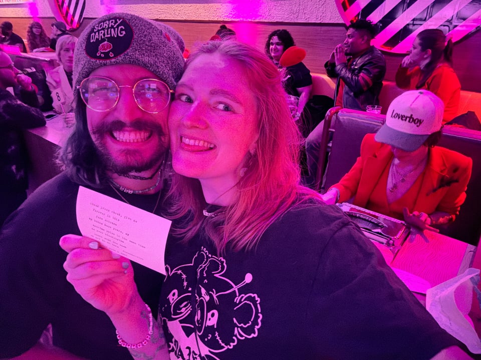 beth and dan, awash in pink light, smile and hold up a fresh typewriter poem. tal is in the background.