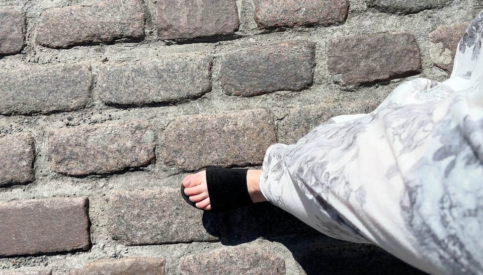 First-person view of a person's foot in a black sandal with a wide strap, striding along cobblestones.