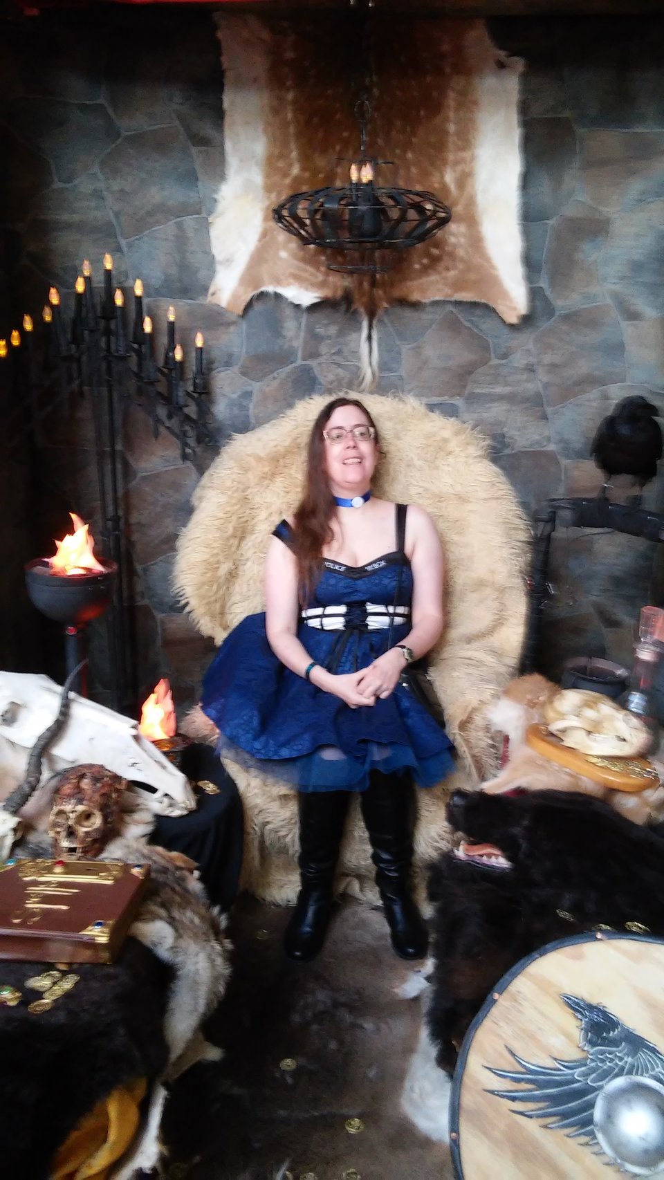 A brunette woman sitting on a chair in a Medieval setting with a wolfskin rug, skull, and book. There is a raven perched to the right of her. She is wearing a TARDIS dress and long black boots.