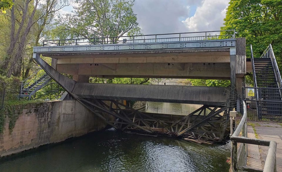 A fairly narrow flood channel. It's spanned by a large plain concrete bridge that is fenced off from public use. Beneath the bridge is a huge concrete and steel rocker with a barrier on one side and a counterweight on the other. The barrier is sitting in the water, closing the gate.