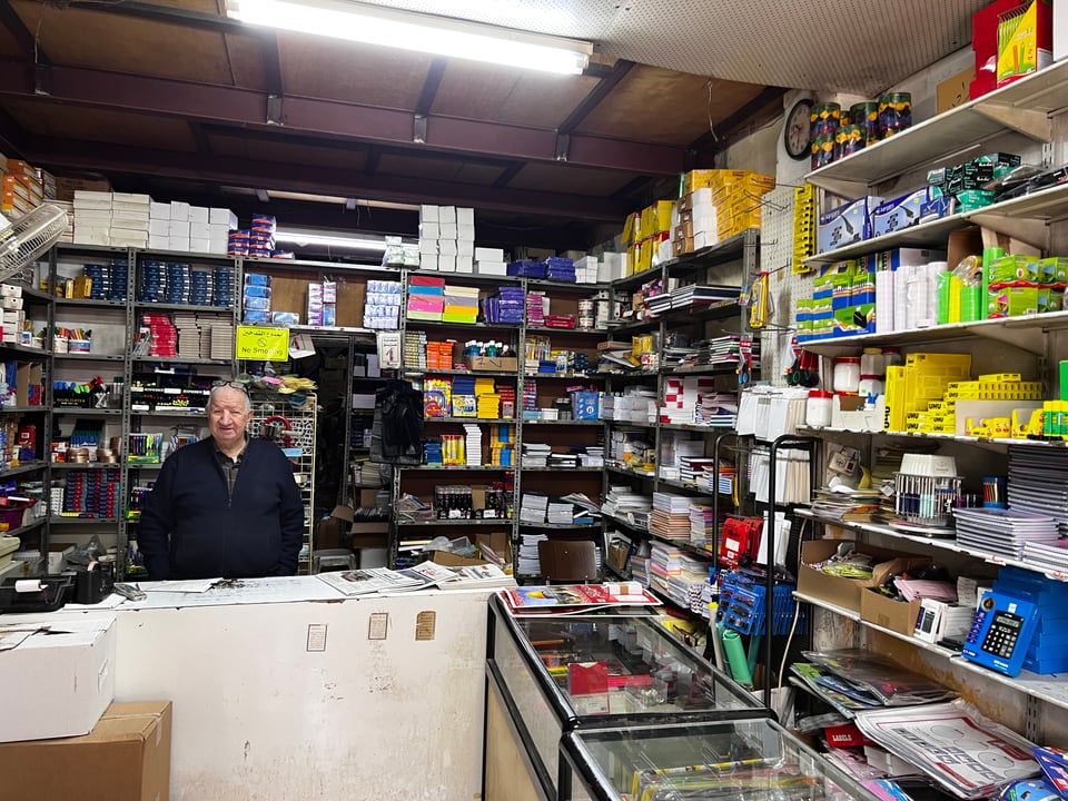 A man standing behind a counter in a store with crowded shelves behind a counter on two sides