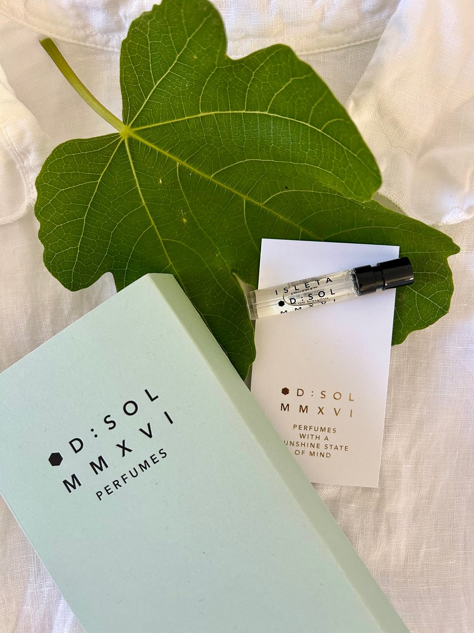 A bright green fresh fig leaf is on a white linen shirt. There's a small glass vial with Isleta by D:SOL Perfumes in. There's a pale blue box with D:SOL branding on and a white and gold business card with the branding in gold. Image by Rowan Ambrose.