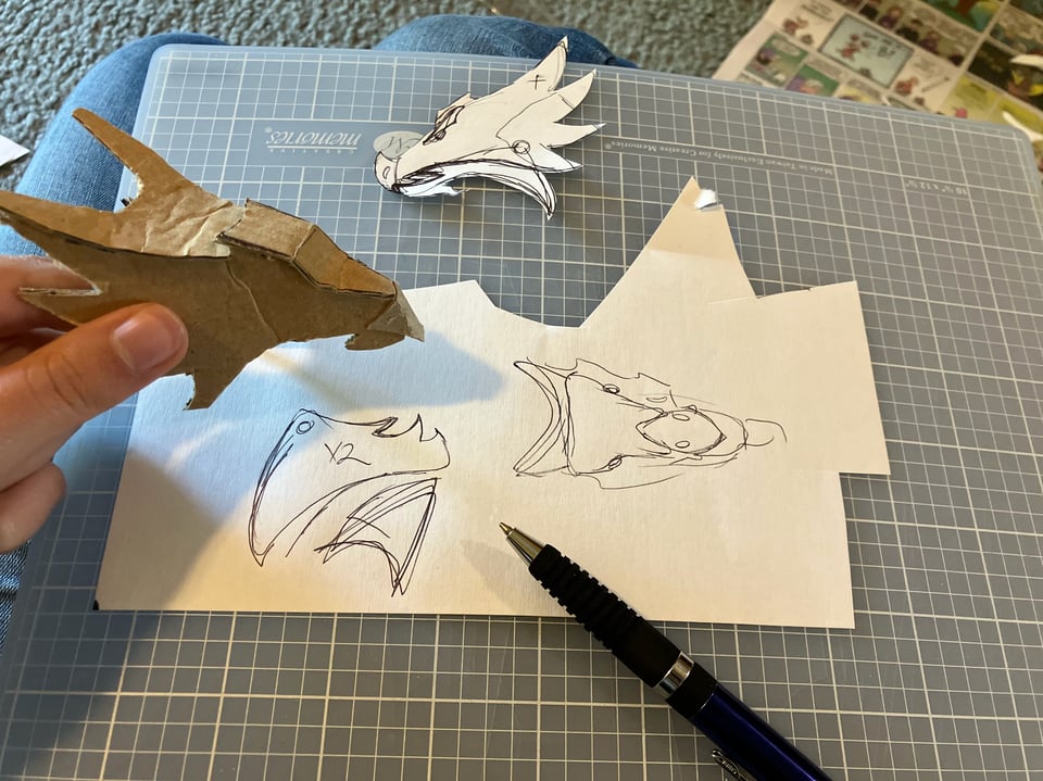 Cardboard dragon head, sans jaw, held next to sketched plans for the head and a pen.